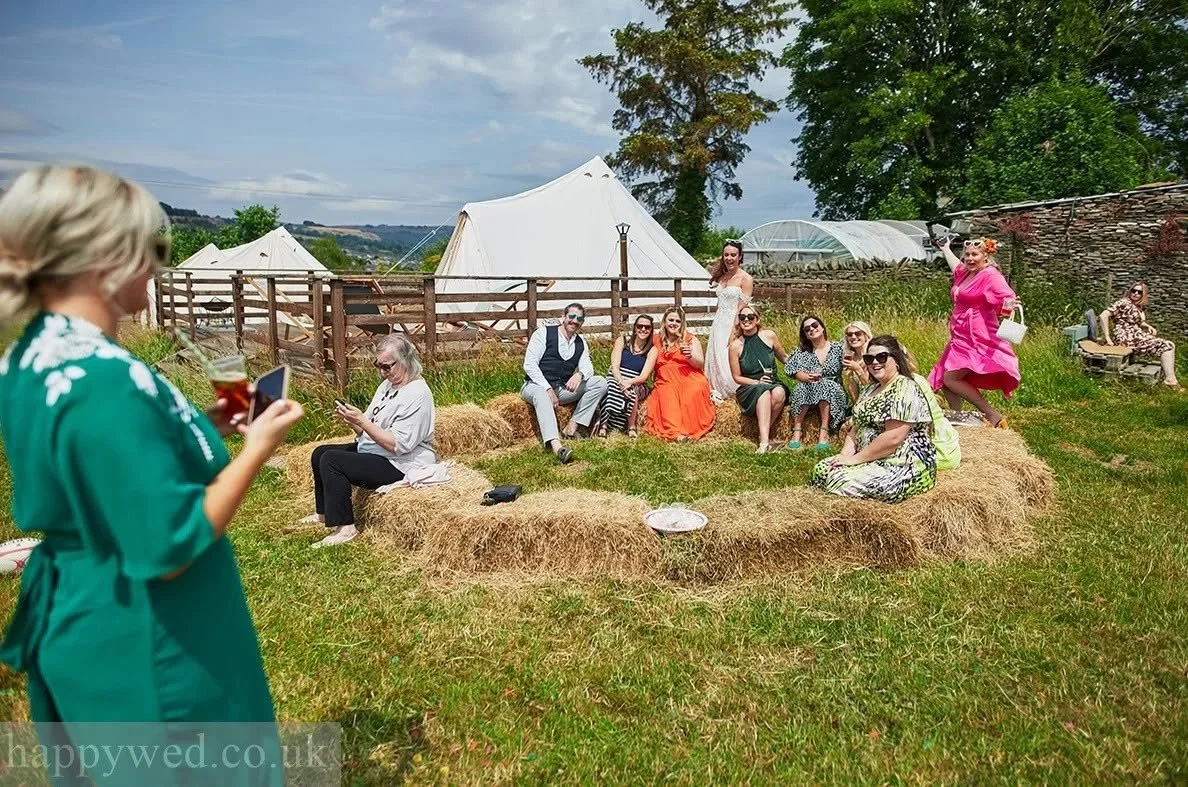 A group of women sitting on haybales in an outdoor setting, with some standing and socializing. Tents are visible in the background, along with trees and a fence. It appears to be a casual outdoor gathering or celebration.