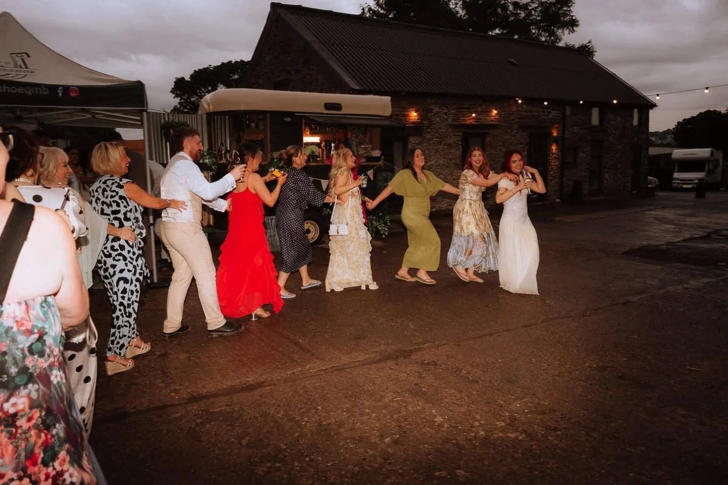 People dancing in a line at an outdoor wedding reception during the evening, with a rustic building and string lights in the background.