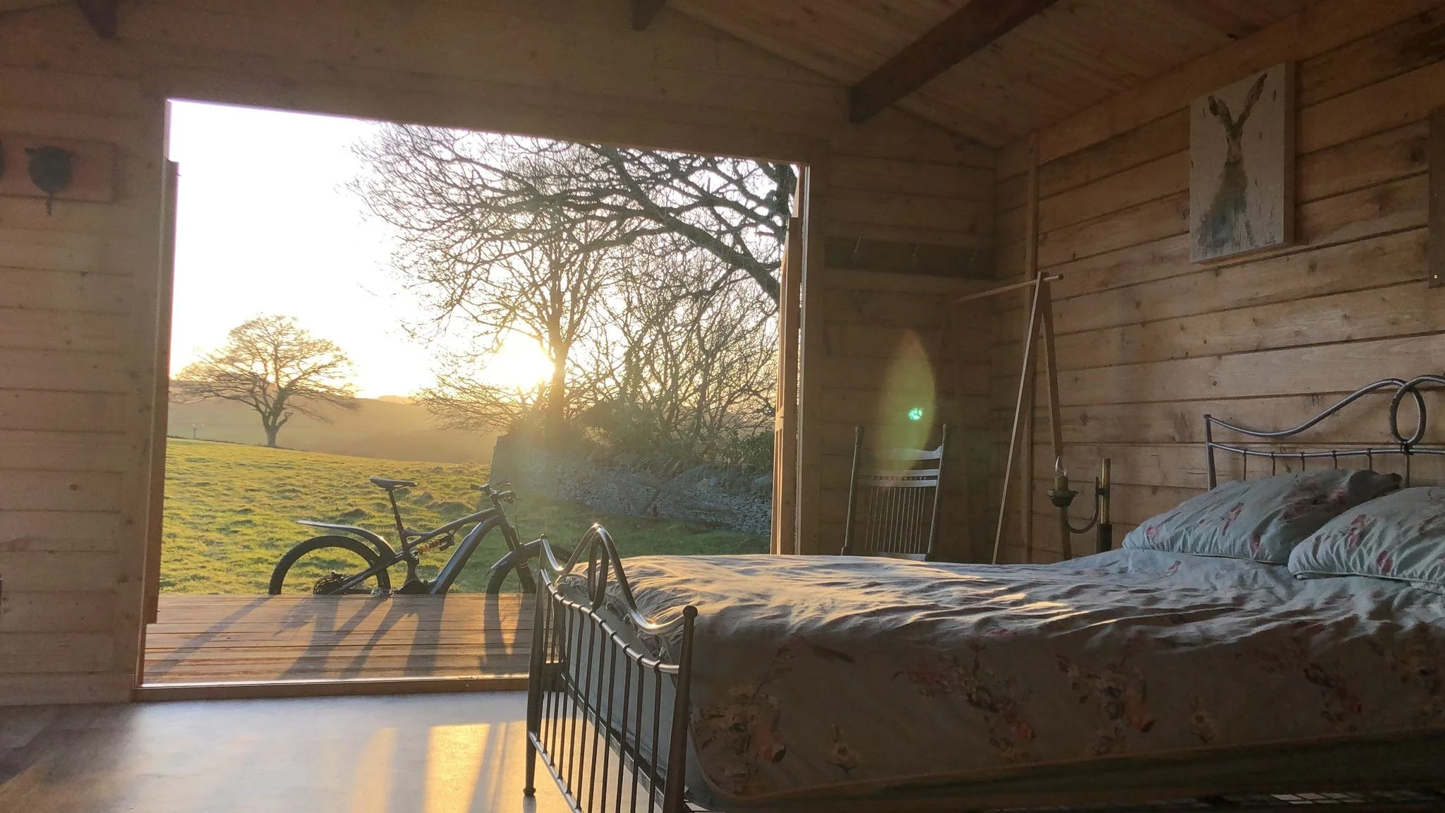 The image shows the interior of a wooden room with an open door revealing a scenic outdoor landscape at sunset, including trees and a grassy field. There are two bicycles and a bed with floral sheets inside the room, along with a small painting on the wall.