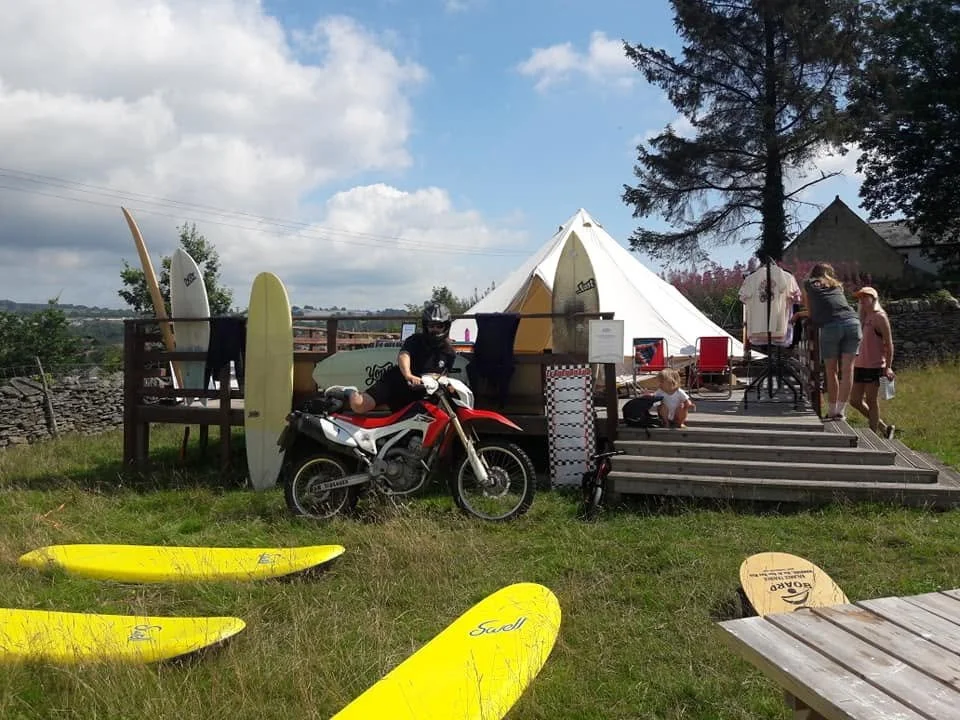A group of people at a beachside or lakeside location with kayaks and a tent, some are preparing or relaxing, with surfboards visible on a deck and grassy area.