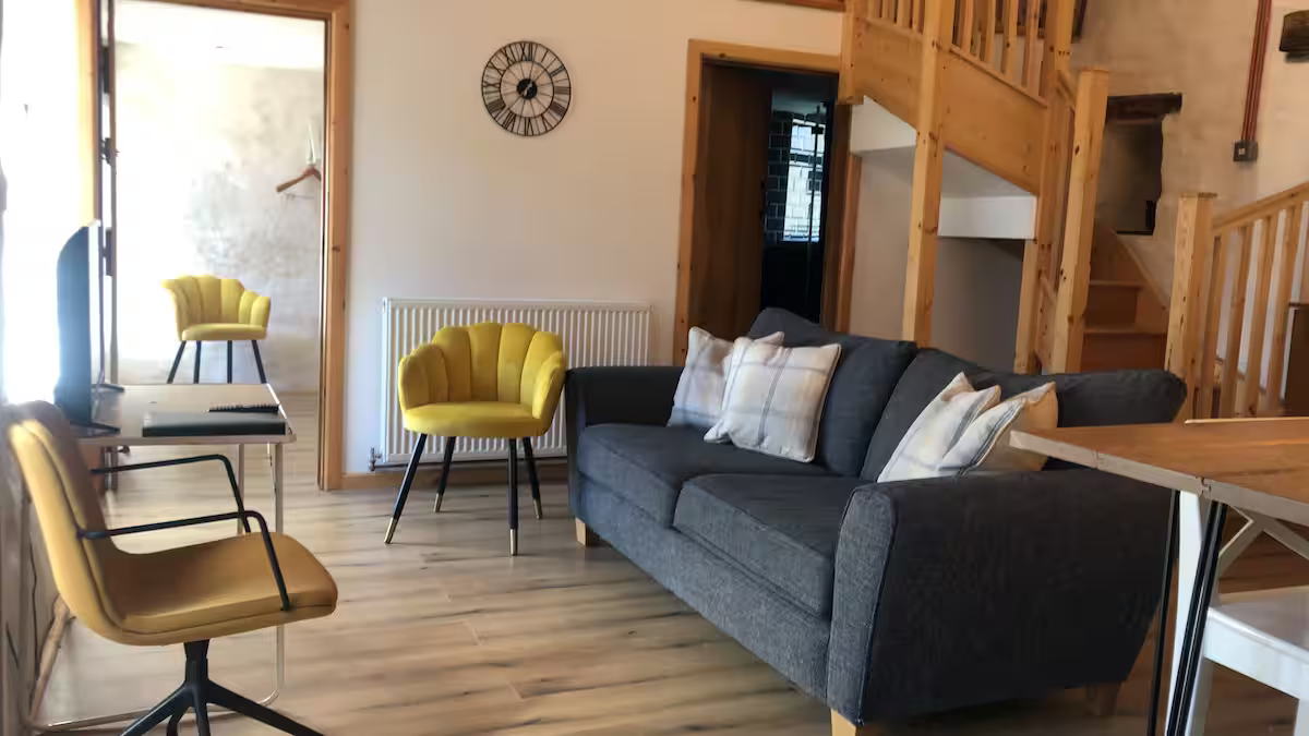 Living room with gray sofa, yellow accent chairs, wooden staircase, clock on white wall, and wooden floors.