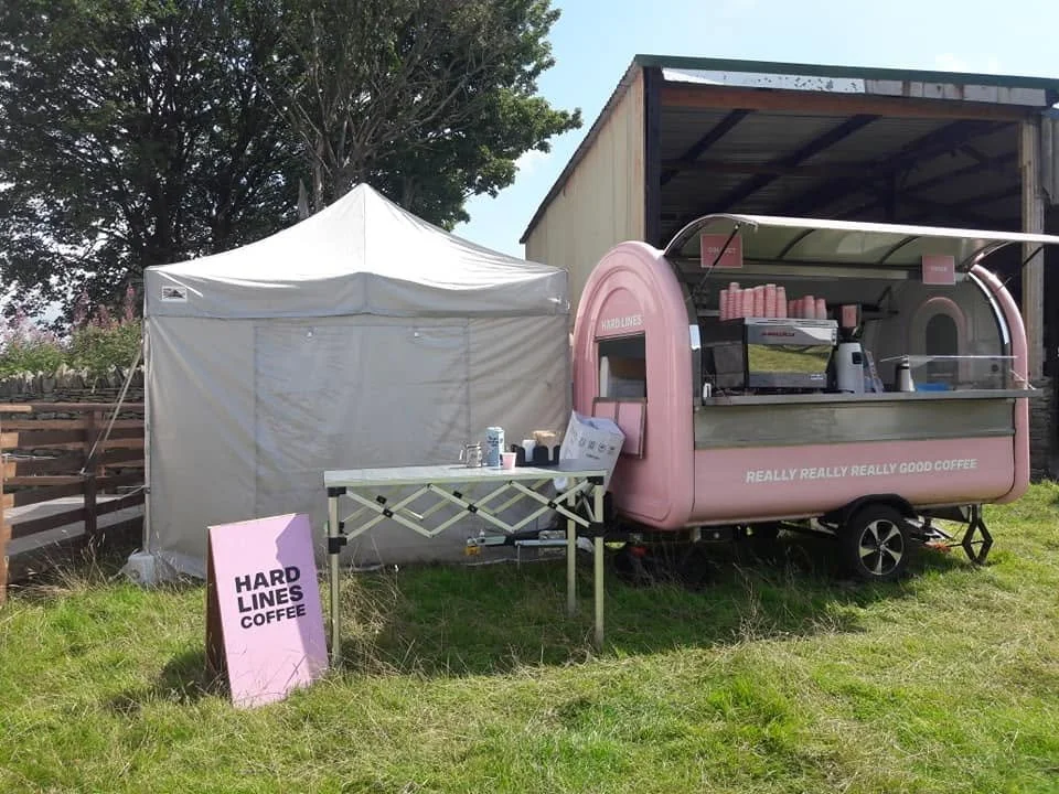 Pink coffee trailer named 'Hard Lines Coffee' and a beige tent set up on grass with a pink sign in front. The trailer has a shelf with pink cups and a coffee machine, with the slogan 'Really Really Really Good Coffee' on it.