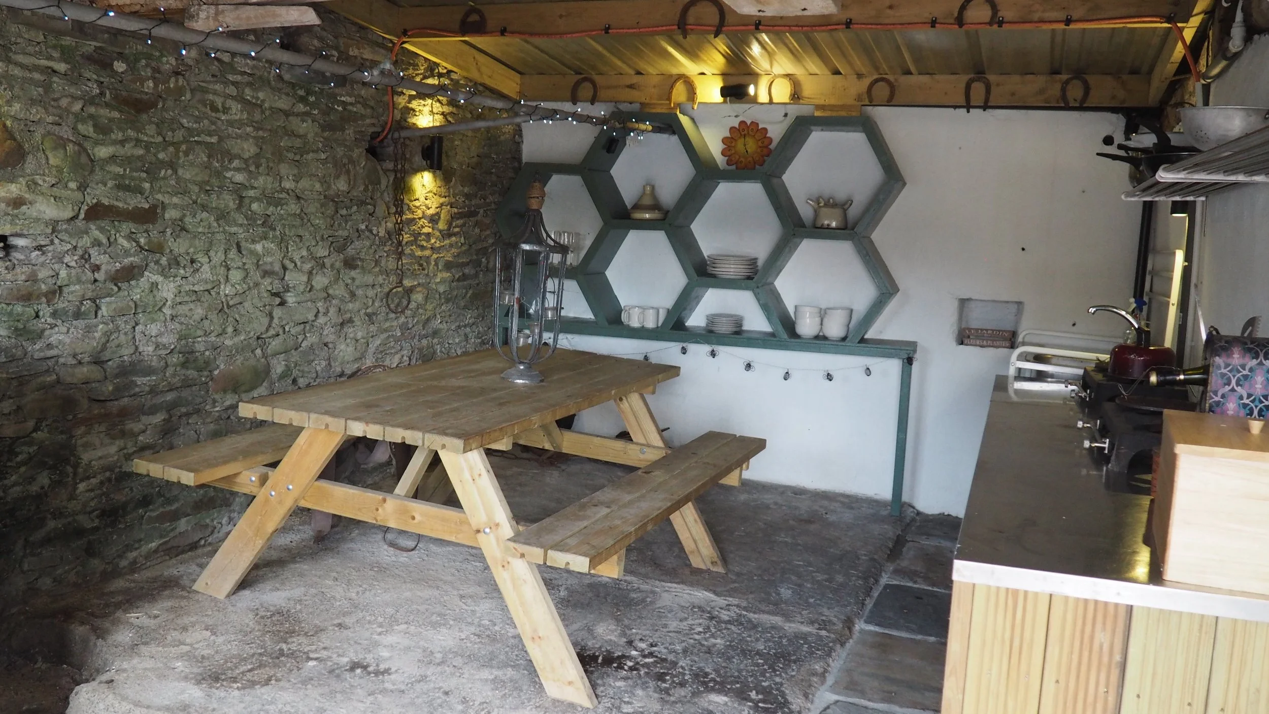 A rustic indoor space with a stone wall, wooden ceiling, and warm lighting. There is a wooden picnic table with attached benches, a shelving unit with plates and cups, and a countertop with various kitchen appliances and utensils.