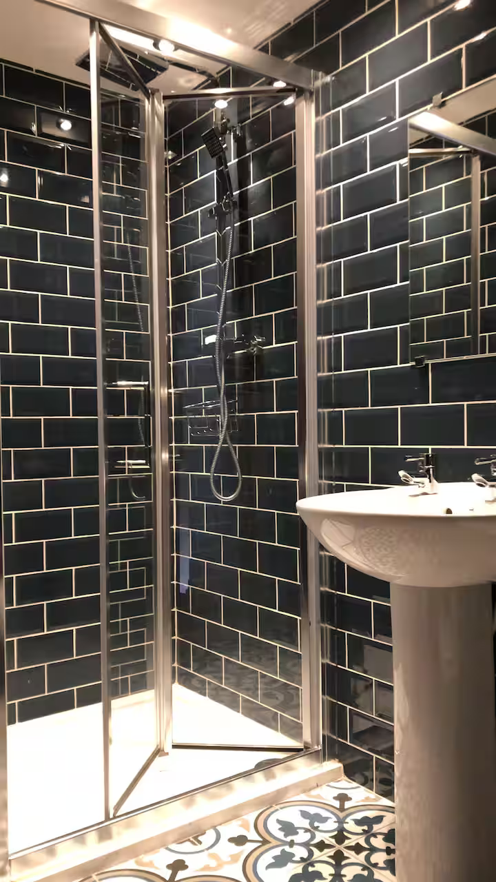 Small bathroom with black tile walls, a glass shower enclosure with a showerhead and sliding doors, and a pedestal sink next to the shower.