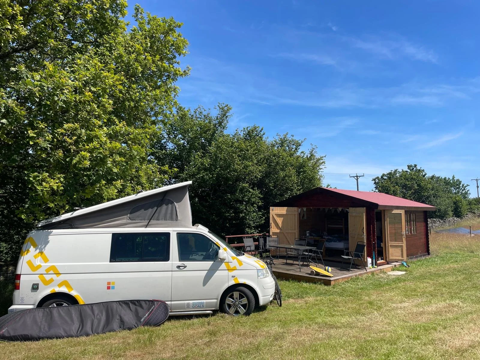 A summer scene with a white camper van parked on grass beside a wooden cabin with open doors, outdoor chairs, a table, and a kayak, surrounded by trees with a clear blue sky in the background.