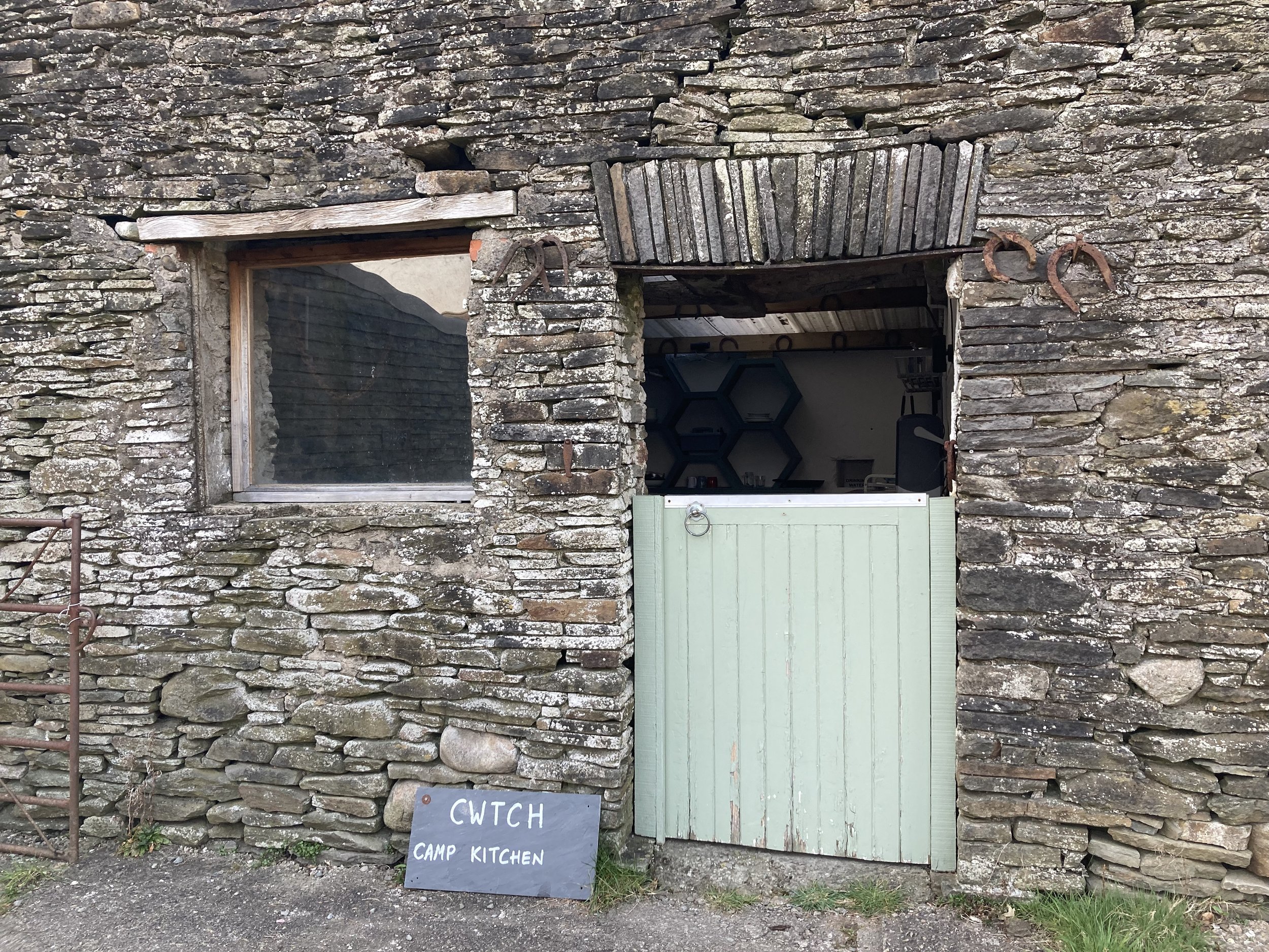 Rustic stone wall with a window and an open doorway, showing a rustic interior with shelves and a chalkboard sign that reads 'CWTCH CAMP KITCHEN'.