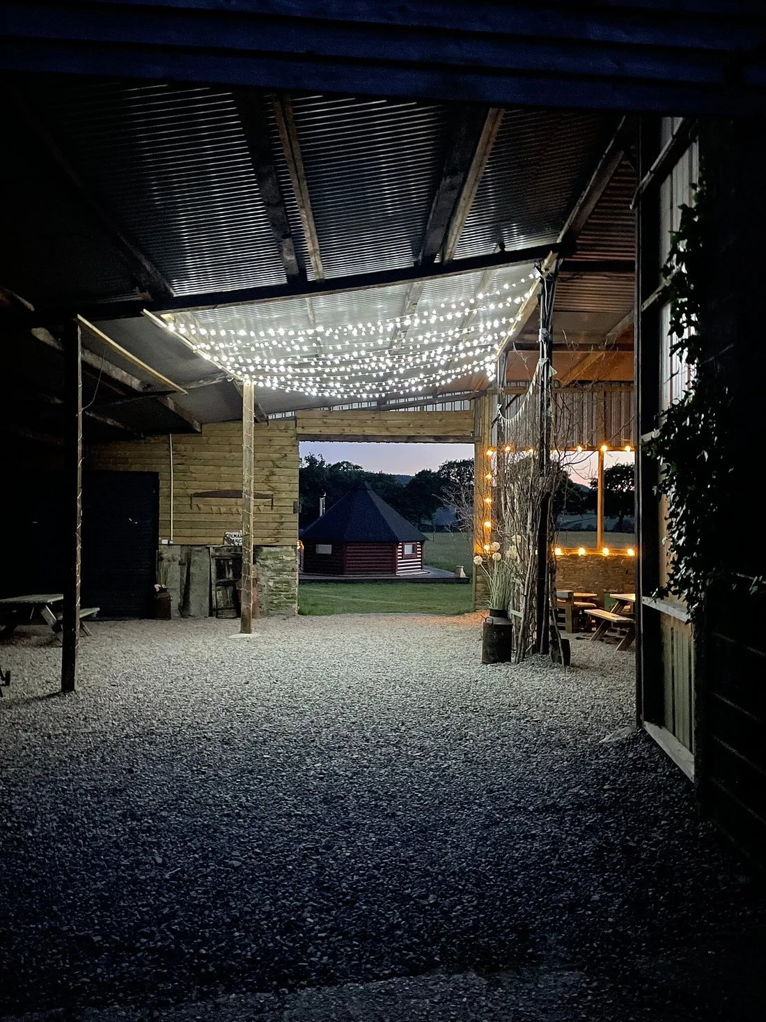 A covered outdoor space decorated with strings of white fairy lights, with a view of a small round building and trees in the background at dusk. Camping