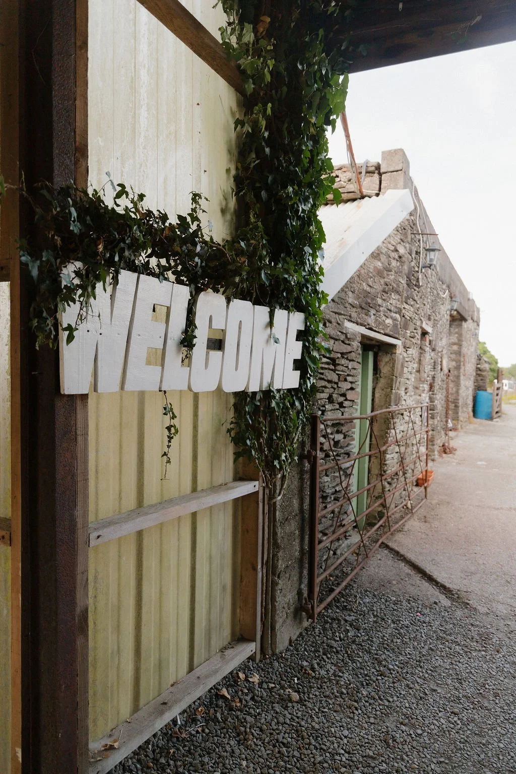 A rustic outdoor scene with a wooden sign reading 'WELCOME' decorated with green vines, set against a weathered wall beside a stone building with a partially open door and a metal railing.