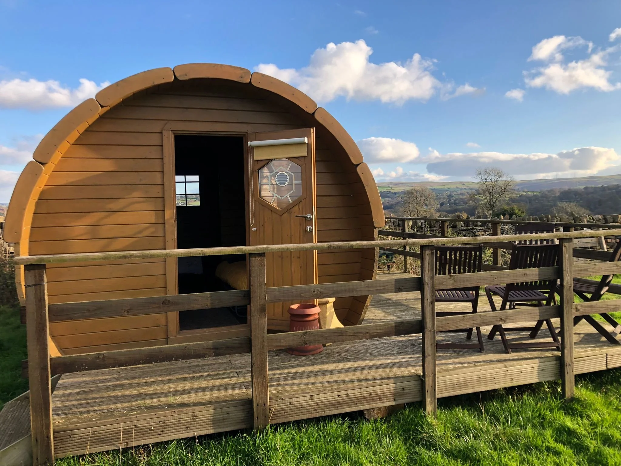 A small wooden pod with a rounded roof and an open door, set on a deck with wooden railings, overlooking a countryside landscape with trees and hills under a partly cloudy sky.