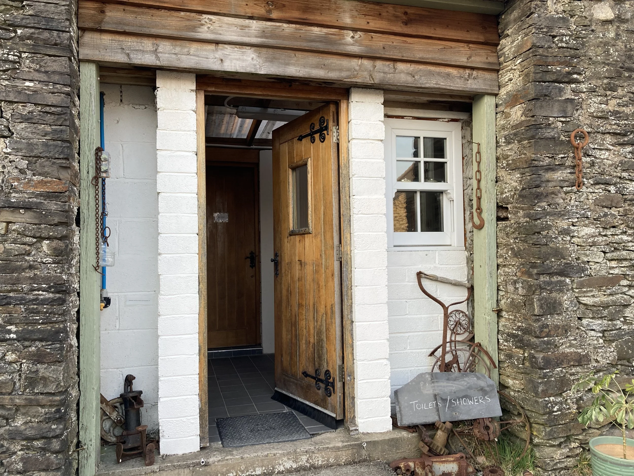 A rustic entrance to public toilets or showers with an open wooden door, surrounded by stone and white brick walls. There is a small black signboard on the ground to the right, labeled "TOILETS / SHOWERS". Various vintage metal tools and ornaments ar