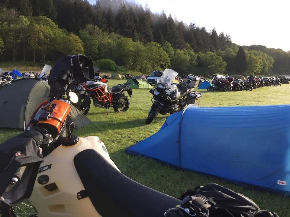 Camping area with motorcycles and tents, set against a backdrop of forested hills.
