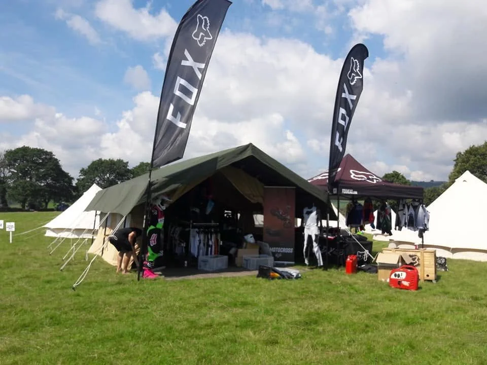 Outdoor event booth with banners, tents, and merchandise, on a grassy field with trees and cloudy sky in the background.