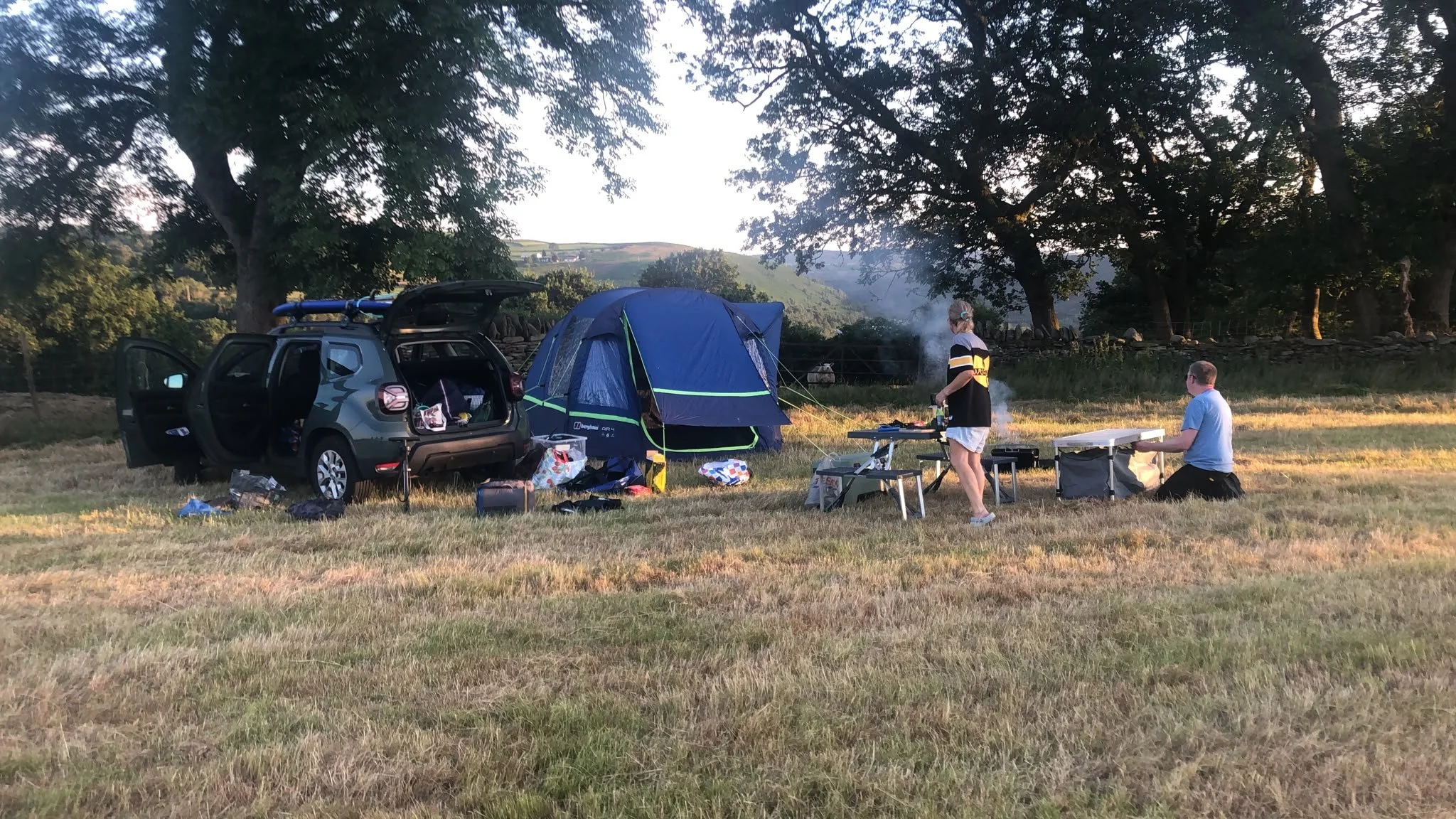 A couple of people camping in a field with a car and a blue tent, surrounded by trees and hilly landscape, during sunset or early evening.