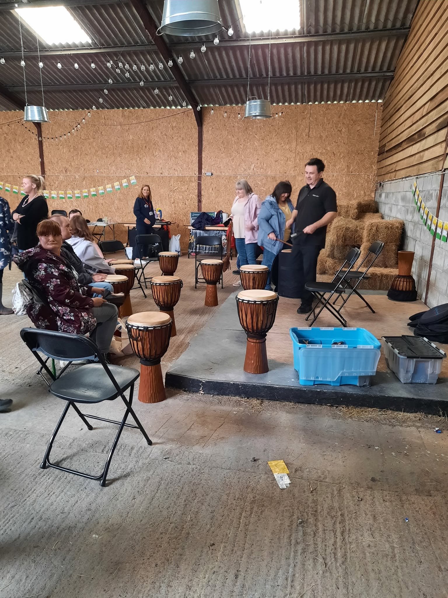People at a college or community center gathering. Some are sitting, some are standing, and one person is playing a drum. The setting has hay bales, grain, and string lights.