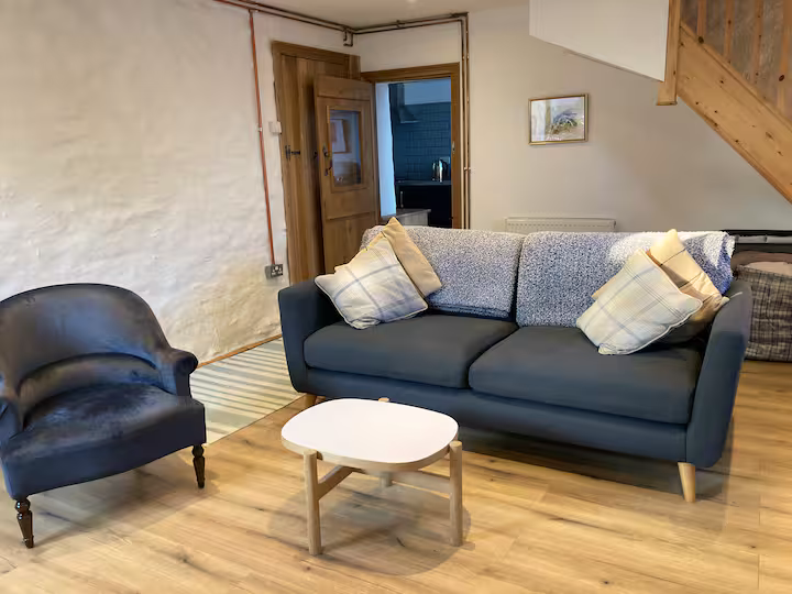 Living room with a gray sofa with multiple pillows, a black armchair, a small white coffee table, wooden staircase, and a door leading to another room.