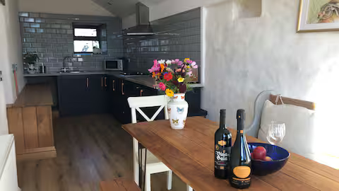 A kitchen and dining area with a wooden table holding a vase of colorful flowers, bottles of wine, a glass of wine, and a bowl of apples; kitchen with black cabinets, gray backsplash, and window in the background.
