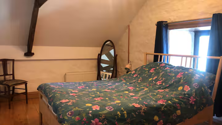 A cozy bedroom with a sloped ceiling, featuring a double bed with floral bedding, a wooden chair, a standing mirror, a window with blue curtains, and a bedside lamp.