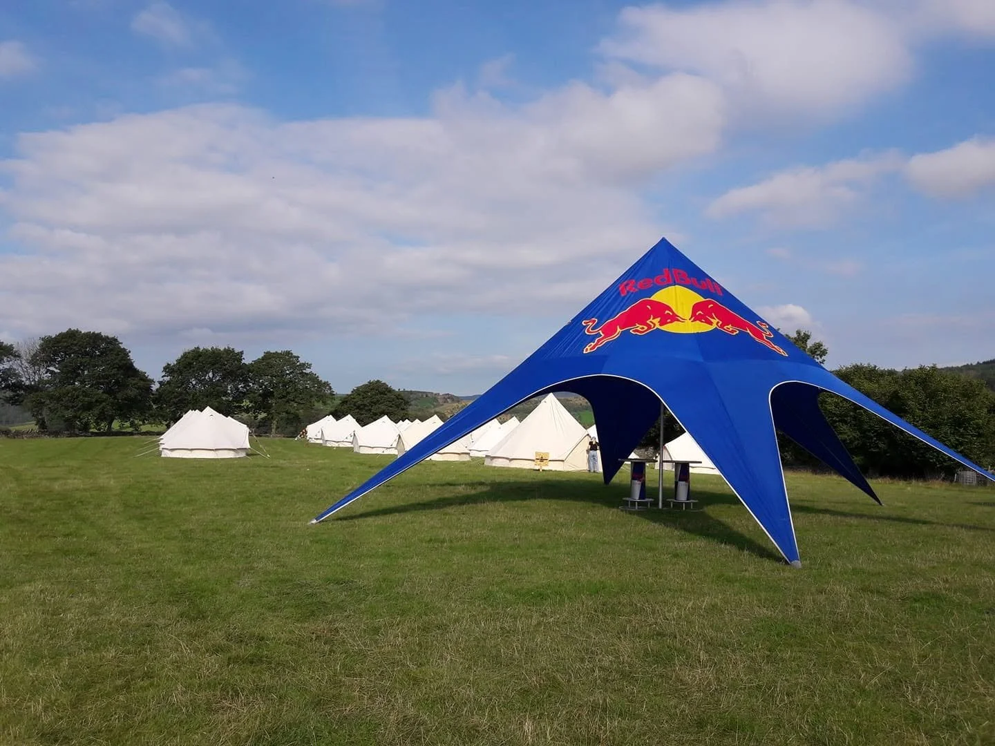 A large blue tent shaped like a star with the Red Bull logo in the center sits on a grassy field, surrounded by smaller white tents and trees under a partly cloudy sky.