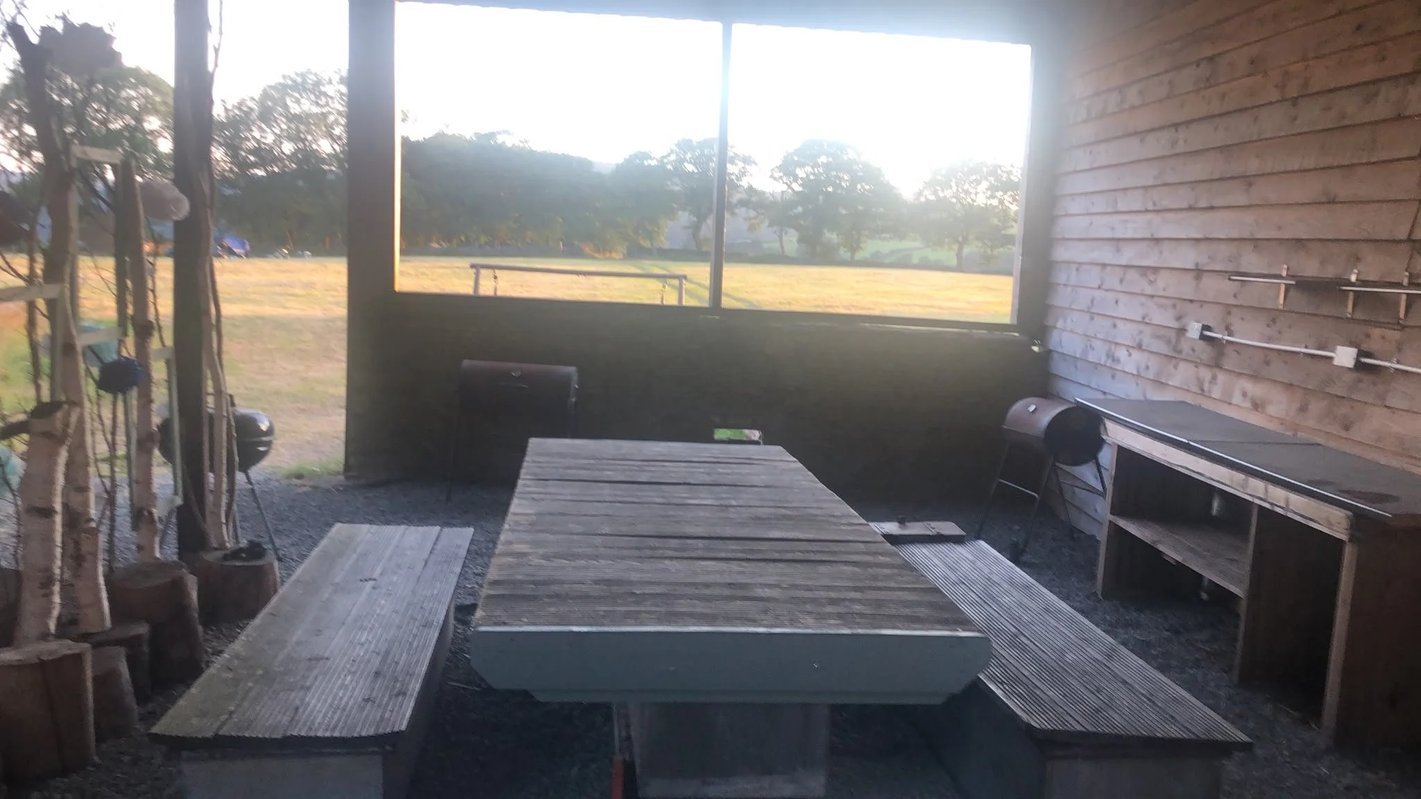 A rustic outdoor patio with a wooden table and benches, a black grill, and a wall with outdoor electrical outlets, overlooking a grassy field with trees in the background.