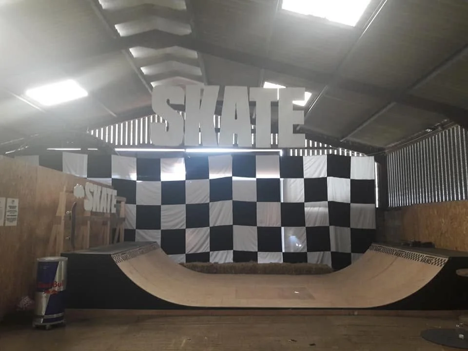 Indoor skateboarding ramp with black and white checkered wall, large white 'SKATE' sign hanging from the ceiling, and a Red Bull cooler to the left.