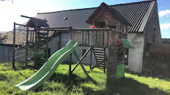 Children's outdoor playset with slide, climbing area, and sandbox, in a backyard with grass and a house in the background.