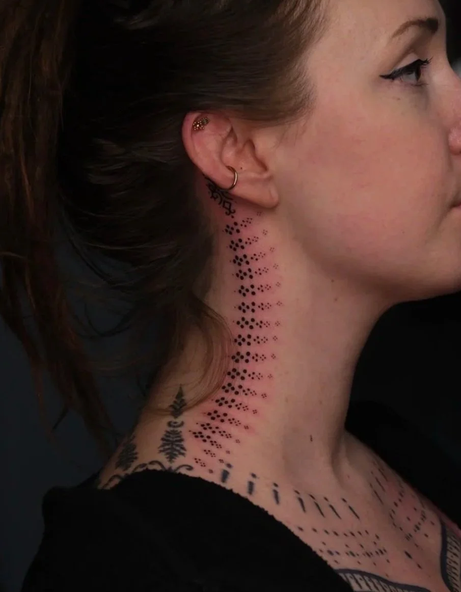 Side profile of a woman with tattoos on her neck and chest, wearing multiple earrings.