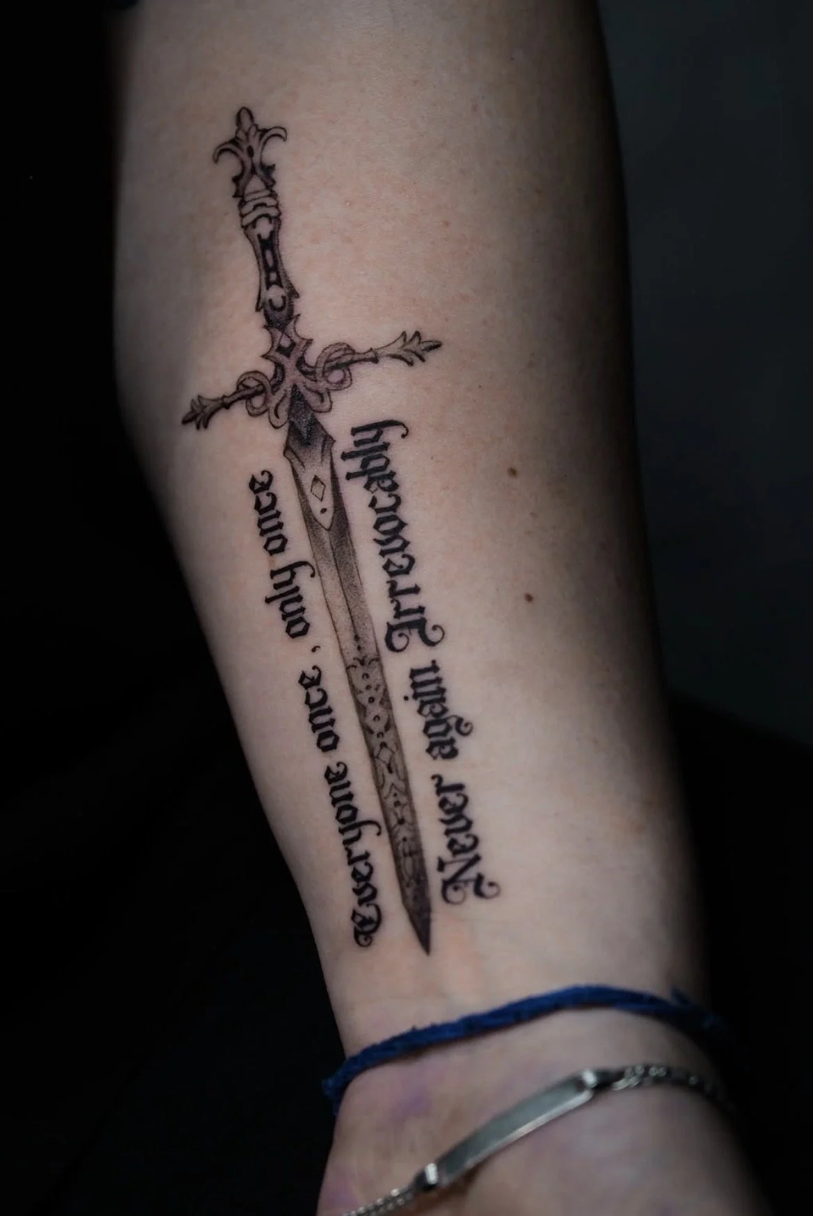 Tattoo of a sword with ornate detailing and a quote written in gothic script, partially visible, on a person's arm.