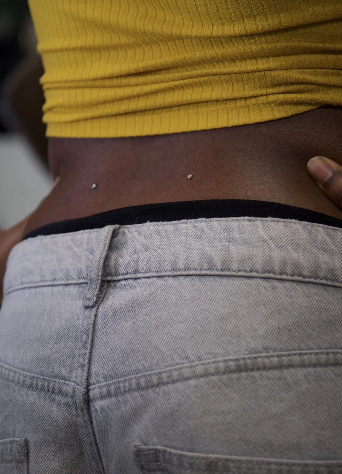Close-up of a person's midsection showing a yellow top, a belly button with a piercing, and gray jeans.
