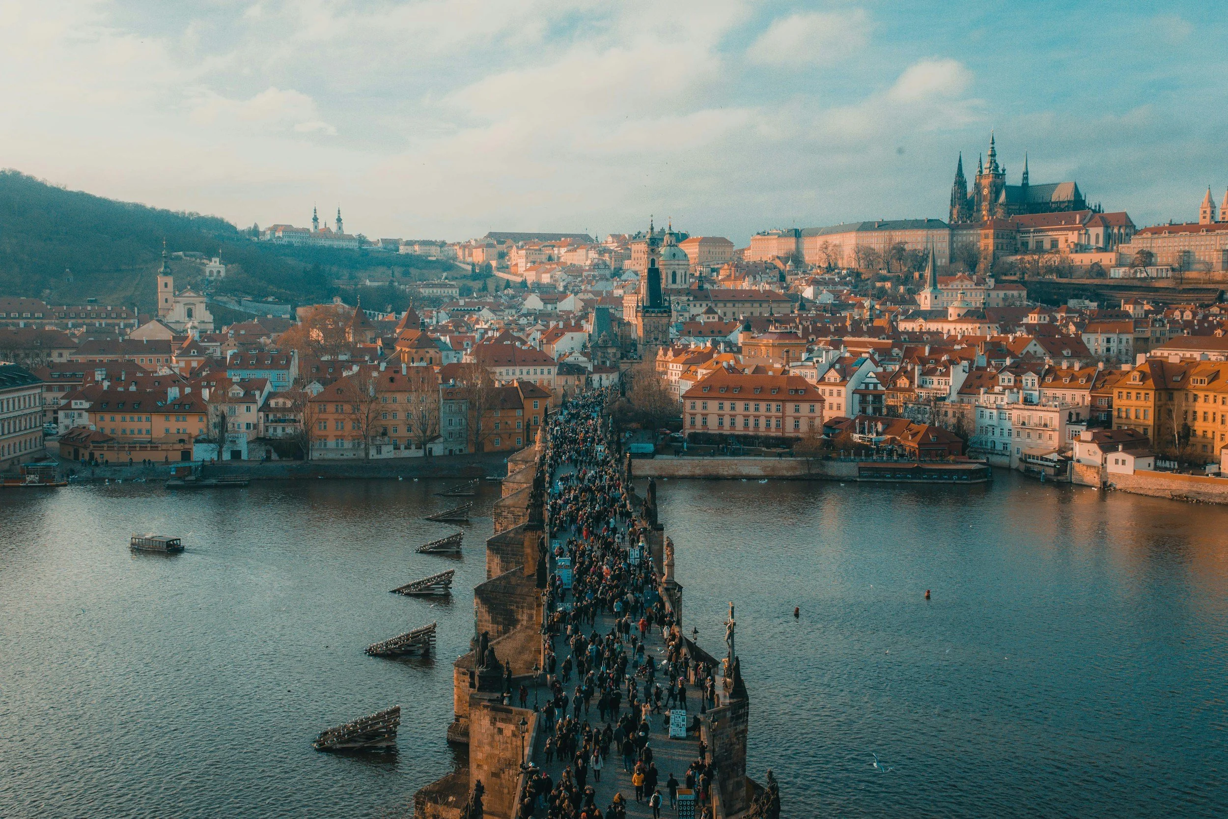 Aerial view of Charles Bridge in Prague, crowded with people, spanning the Vltava River. Historic buildings with red rooftops and Prague Castle are visible in the background under a partly cloudy sky.