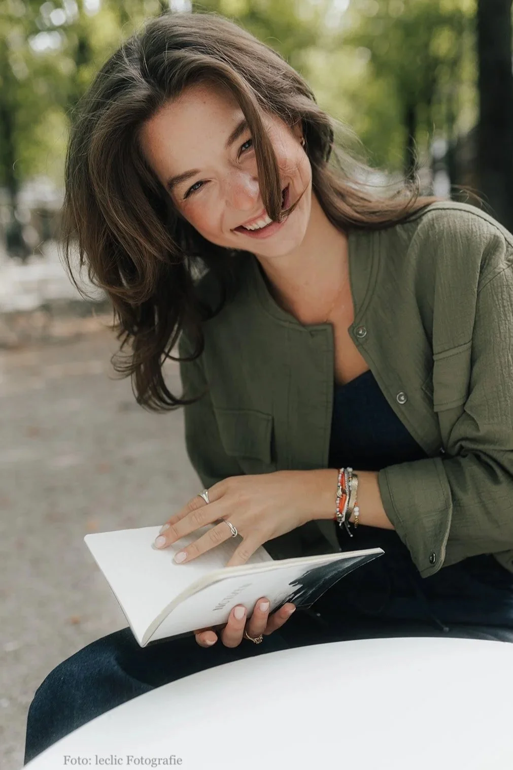 Eine lachende junge Frau mit braunen Haaren liest ein Buch im Freien, umgeben von Bäumen.