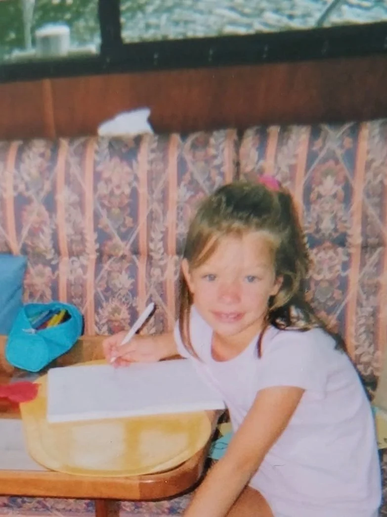 Ein kleines Mädchen sitzt an einem Tisch, schreibt auf einem Blatt Papier mit einem Stift, hat braune Haare und trägt ein weißes T-Shirt.