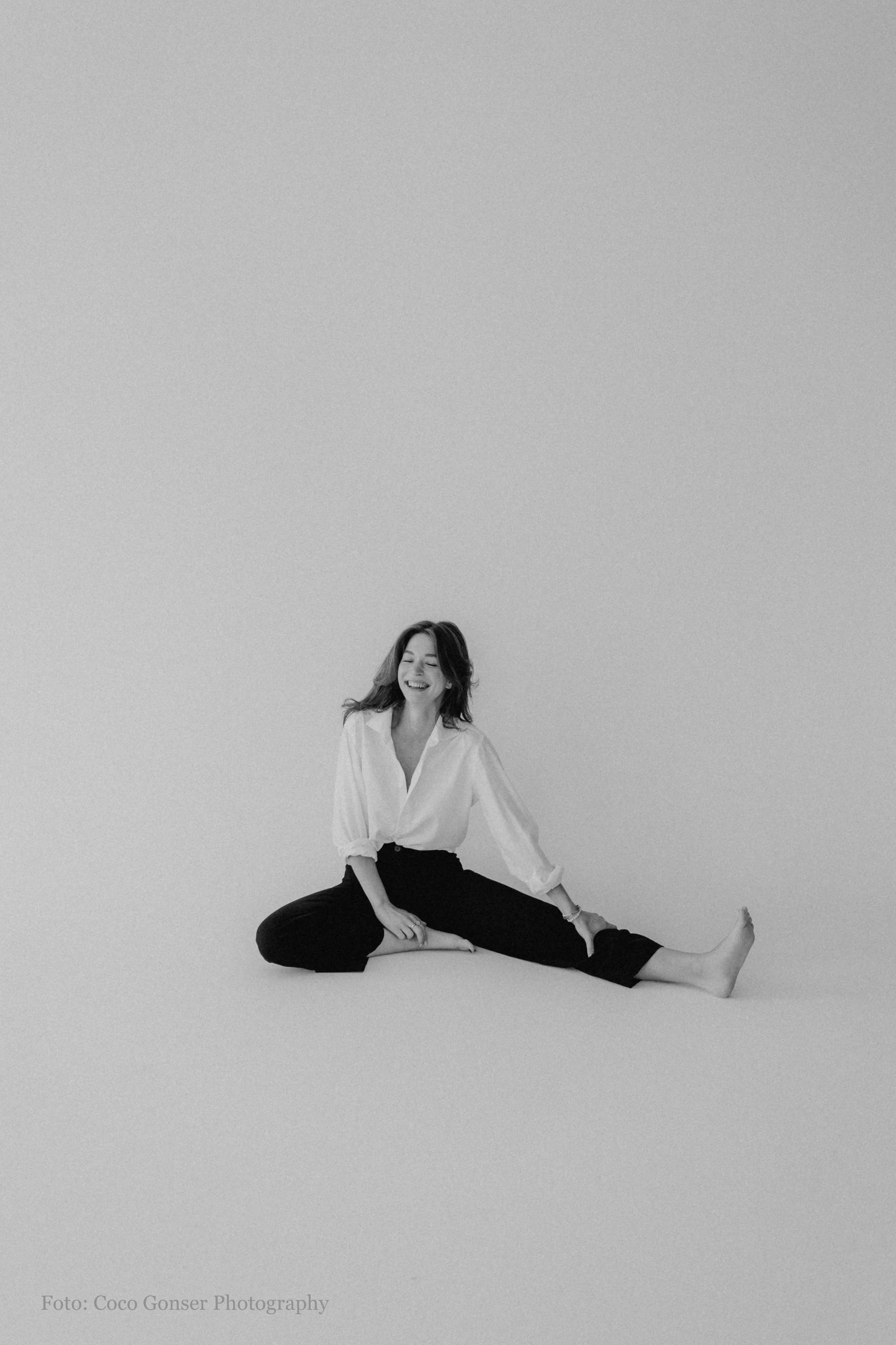 Eine lachende Frau sitzt auf einem Boden, trägt eine weiße Bluse und schwarze Hosen, hintergrundfrei.