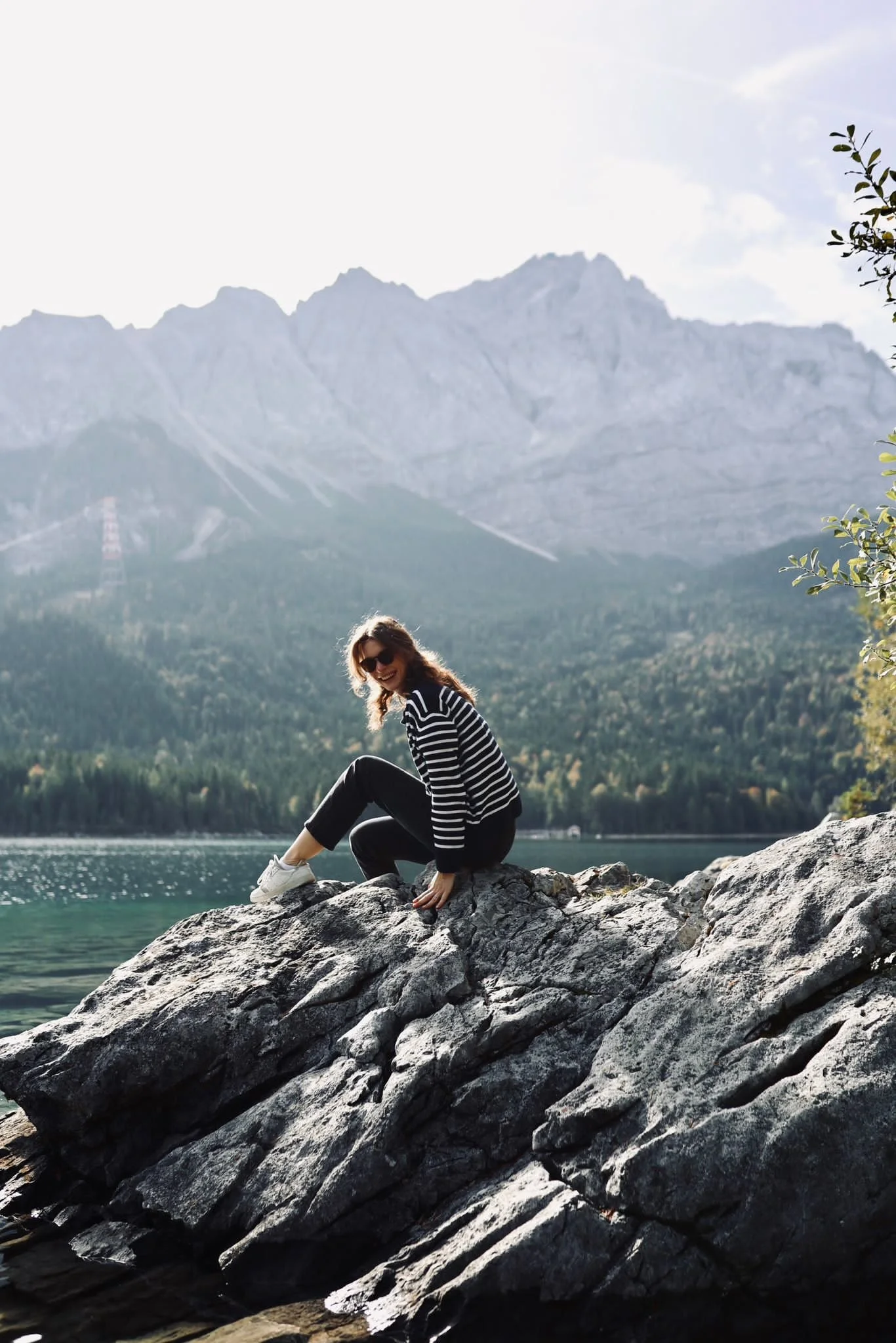 Eine lachende Frau sitzt lachend auf einem großen Felsen am Ufer eines Sees mit Bergen im Hintergrund, tagsüber bei Sonnenschein.