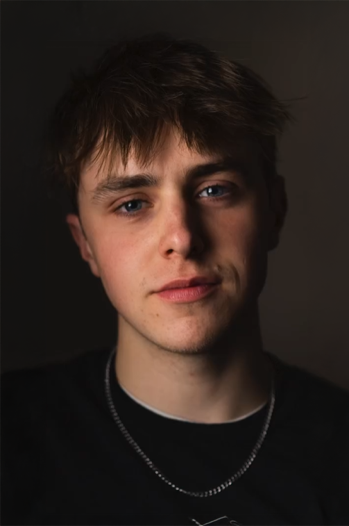 Portrait of a young man with light skin, short brown hair, and blue eyes, wearing a black shirt and a silver chain necklace, against a dark background.