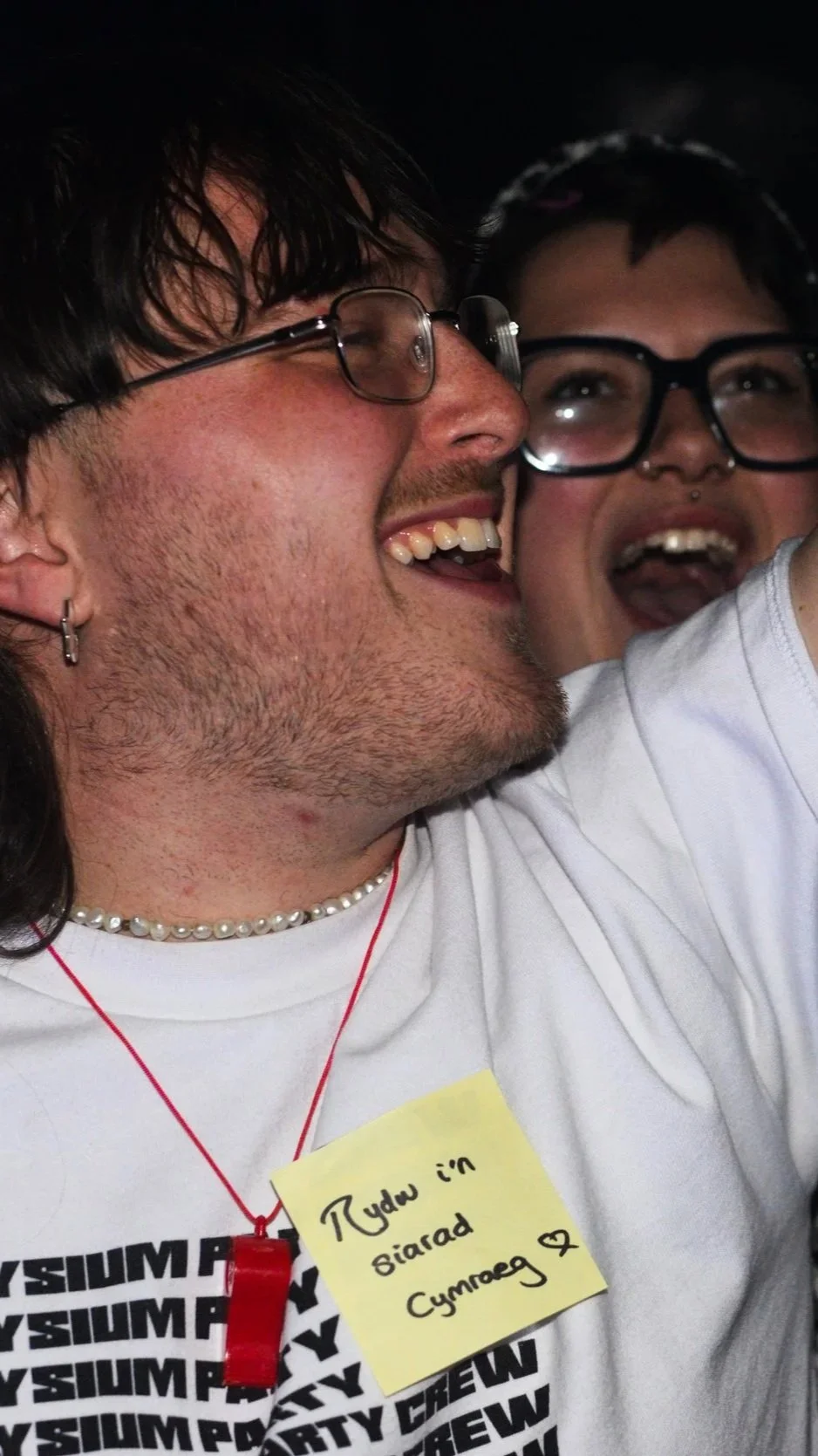 Two people smiling and taking a selfie together, one wearing glasses and a white shirt with a yellow note that says in Welsh, 'I'm tired in Welsh,' with a heart symbol. They are close and joyful.