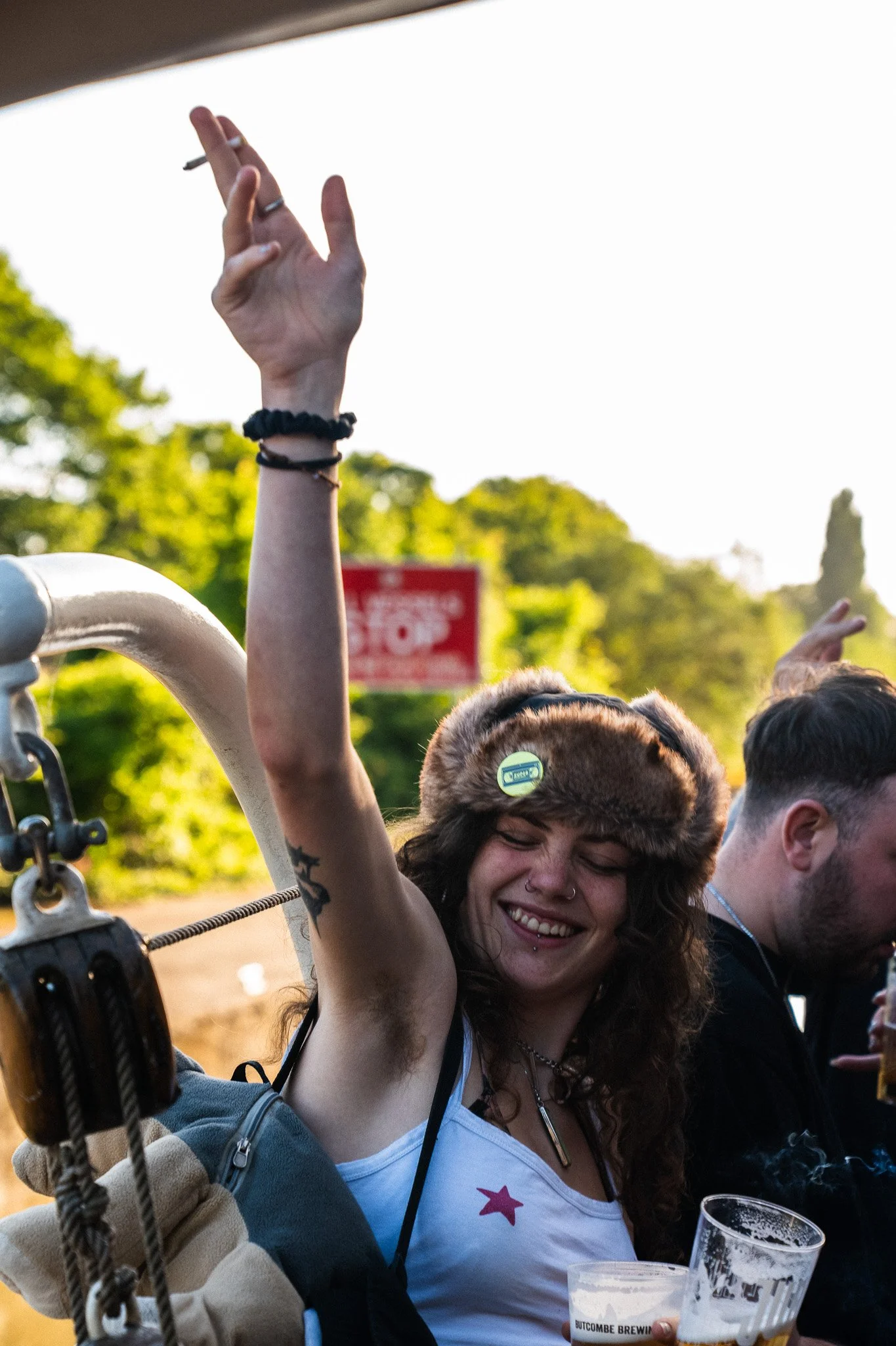 A young woman with curly hair, nose and lip piercings, and a fur hat, smiling and raising her hand making a peace sign at an outdoor gathering, holding a plastic cup of beer.