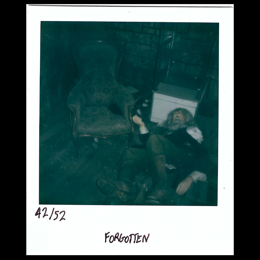 A person with long hair and beard, lying on the floor next to an antique armchair, with a vintage radio or speaker nearby, in a dimly lit setting. The photo is labeled '42/152' and 'FORGOTTEN'.