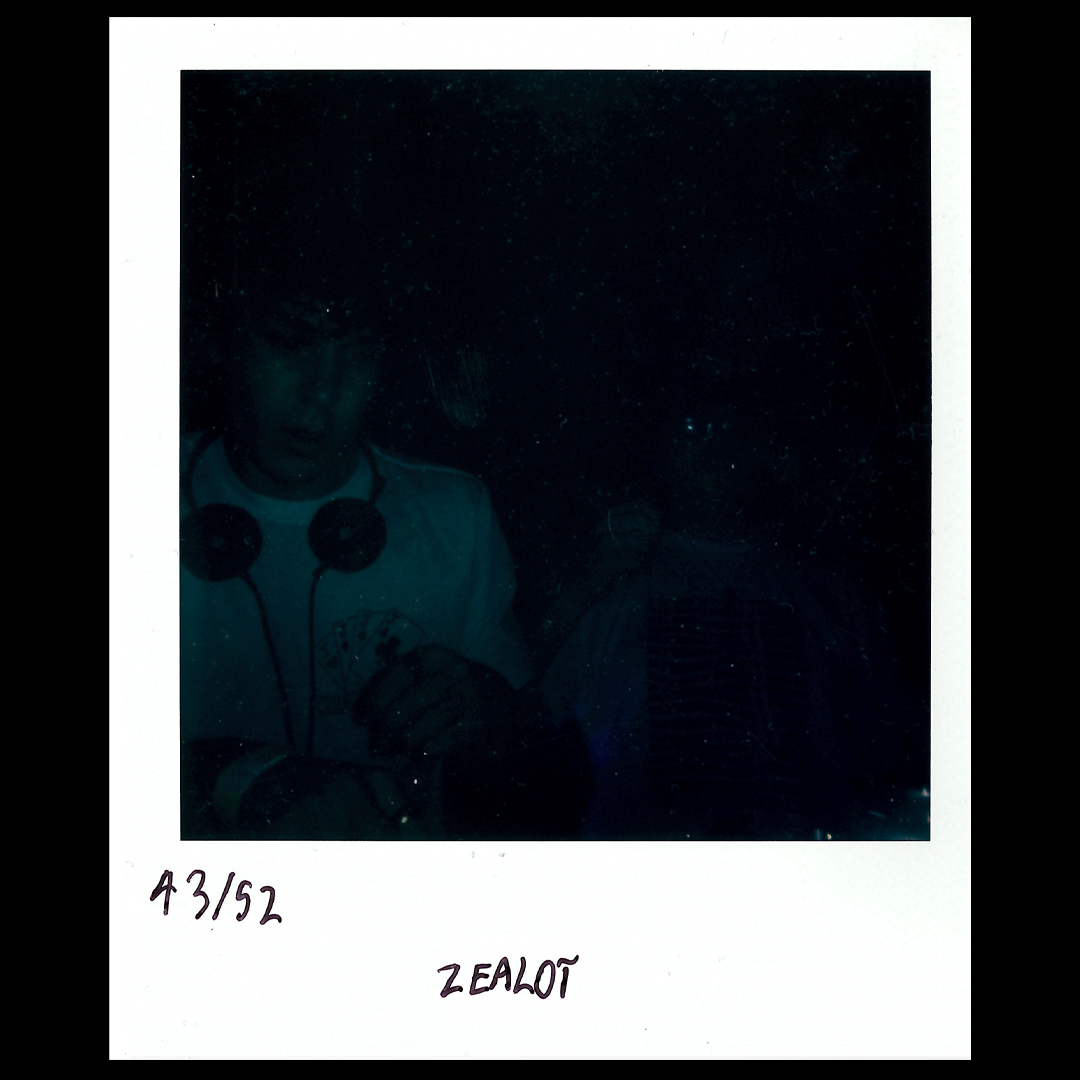 A dark, vintage Polaroid photo showing a person wearing headphones holding playing cards, with the handwritten notes 'A3/52' and 'ZEALOT' at the bottom.