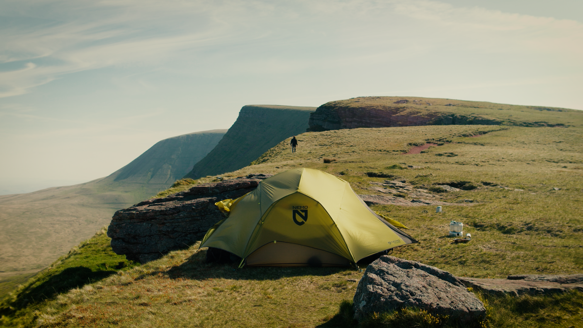 A yellow tent pitched on a grassy hillside with a large rock nearby. In the background, cliffs and a person walking along the hill, under a cloudy sky.