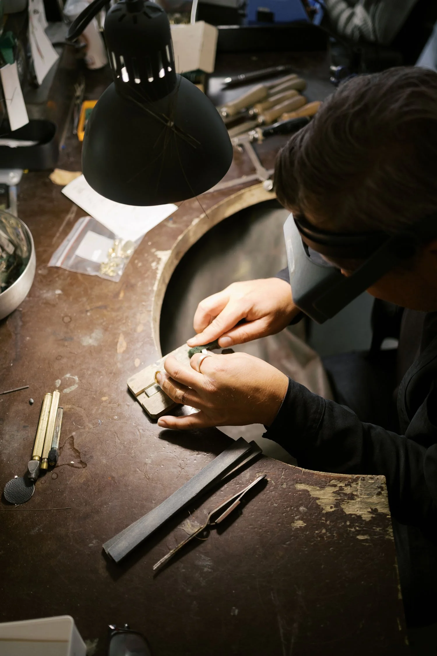 Persoon die met een loep en gereedschap aan een sieraden- of edelsteenwerk werkt onder een lamp. Op de tafel liggen diverse gereedschappen en kleine onderdelen.