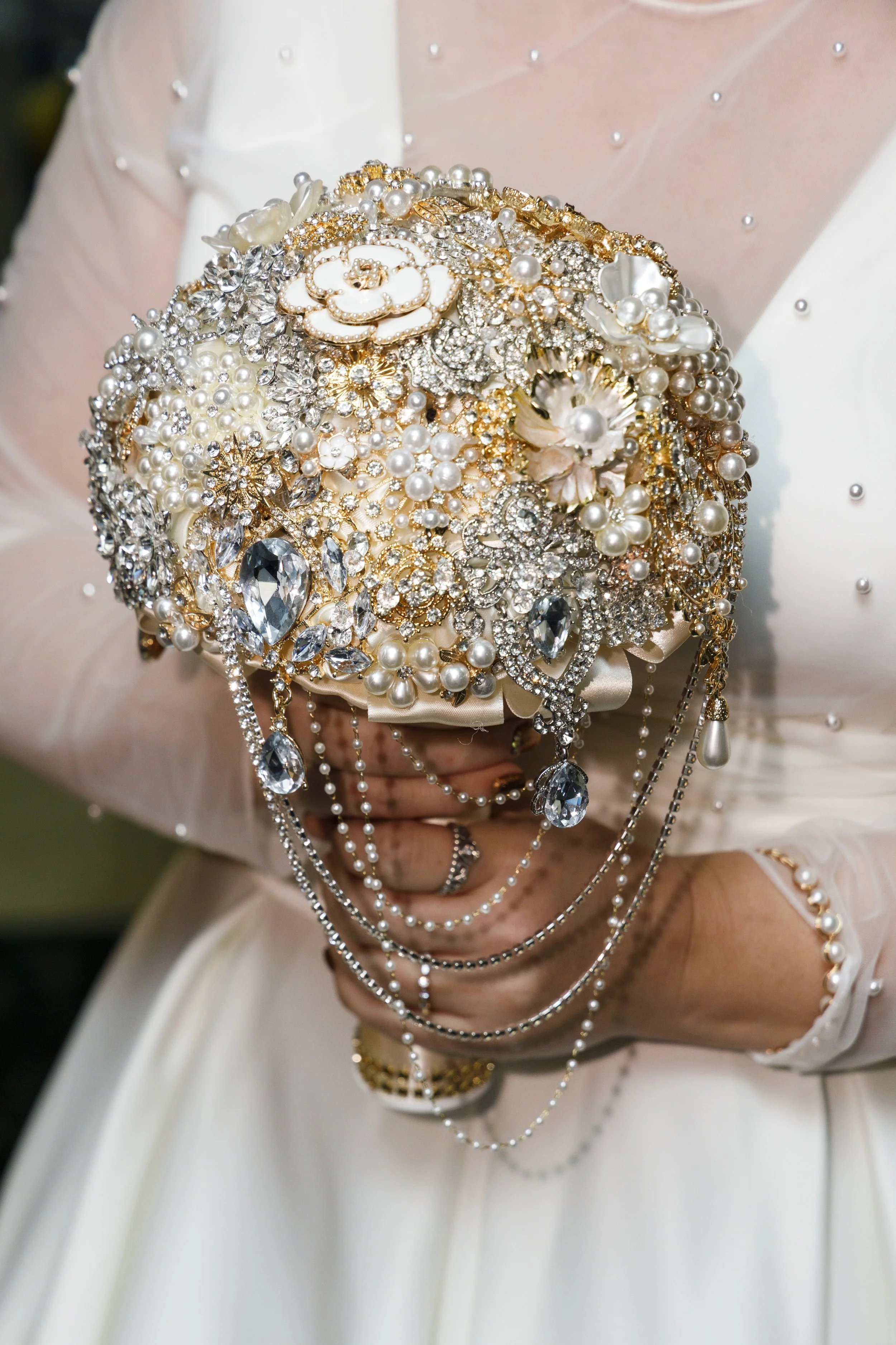 A person holding a bouquet decorated with pearls, rhinestones, and jewelry, wearing a white dress with pearl accents. Now offering the most gorgeous Brooch Bouquets in Wisconsin.