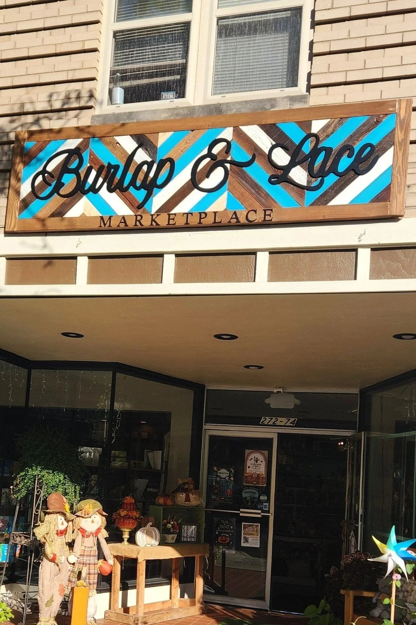 Sign reading 'Burlap & Lace Marketplace' outside a storefront, with some fall decorations and scarecrow figures nearby.