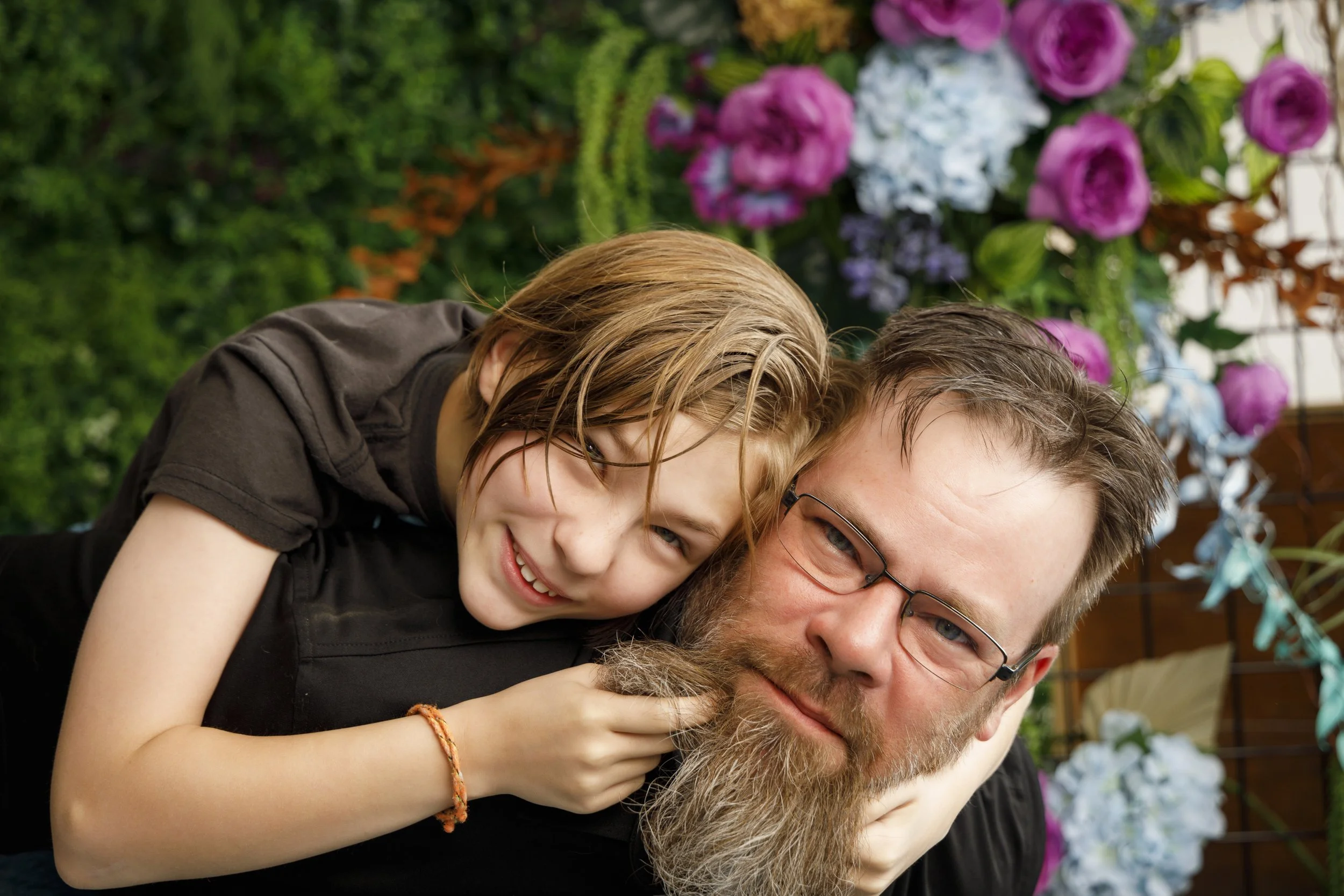 A young girl with wet hair smiling and resting her head on a bearded man's shoulder, both outdoors with colorful flowers in the background.