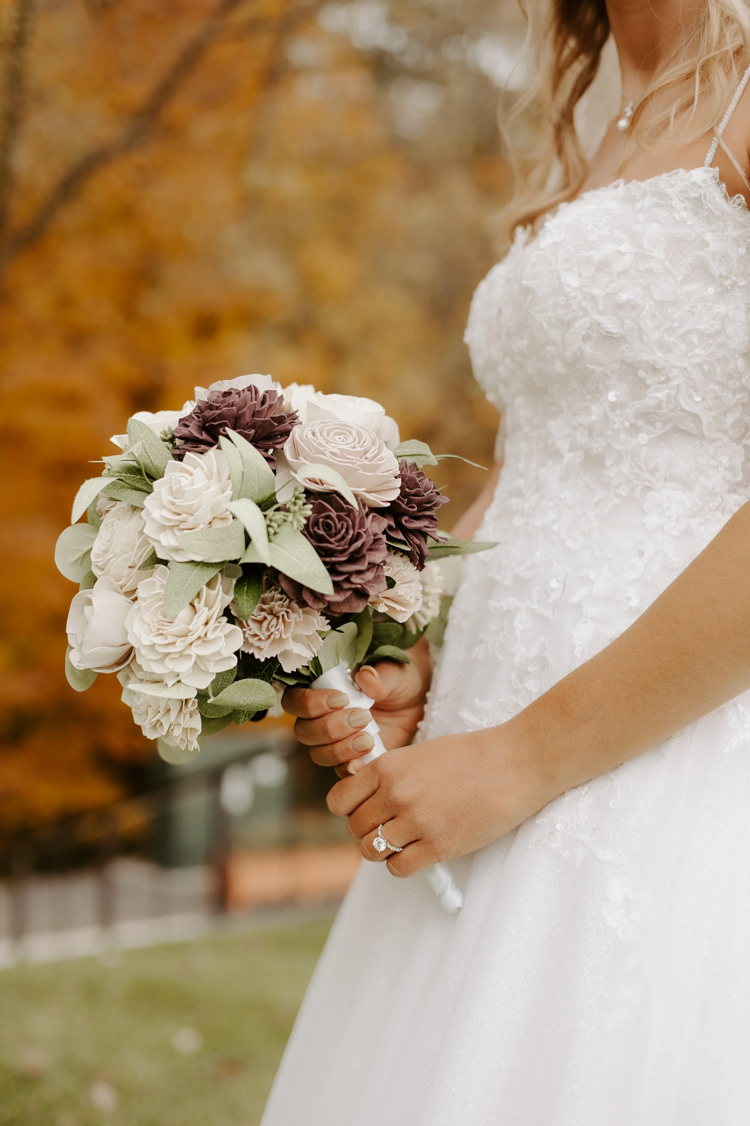 Bride holding a bouquet of purple, white, and cream-colored wooden flowers outdoors with autumn leaves in the background. We specialize in wood flower wedding bouquets and boutonnieres. 