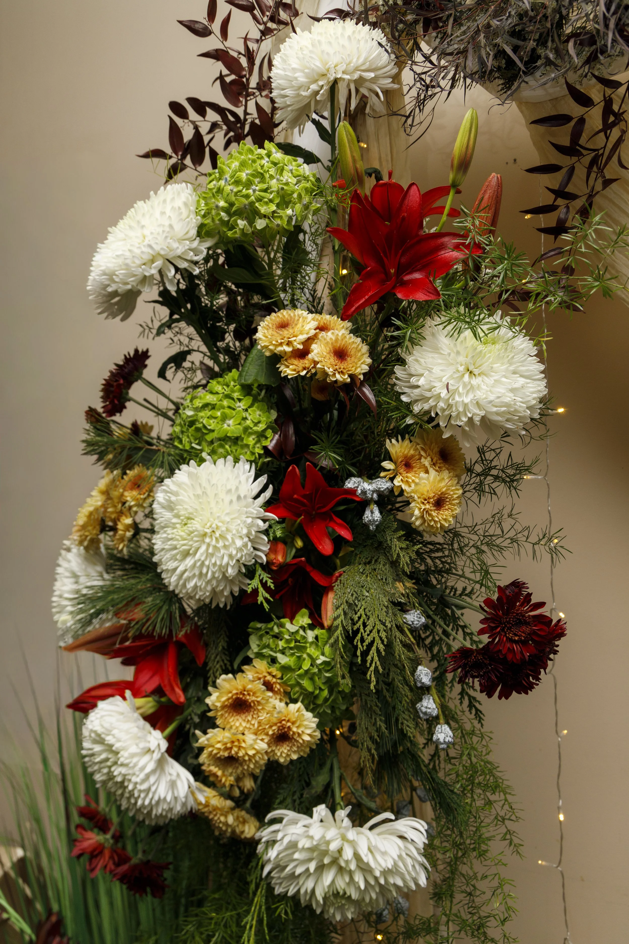Floral Arches make a Statement. A colorful floral arrangement featuring white, yellow, red, and green flowers, with greenery and small decorative ornaments, against a beige background with small string lights.