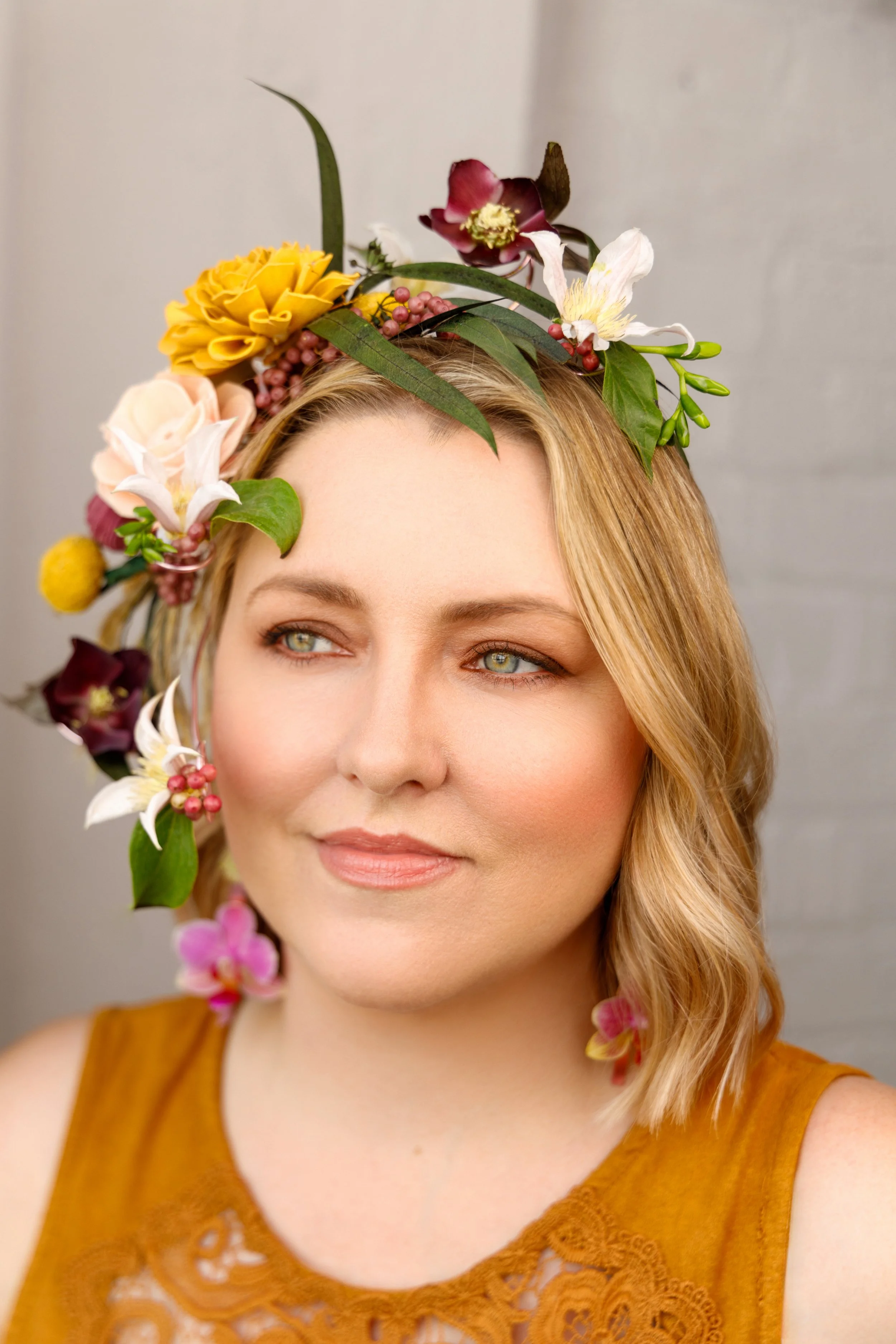 A woman with shoulder-length blonde hair wearing a yellow sleeveless top, adorned with a floral crown made of various colorful flowers and green leaves. We love wearable flowers, flower crowns, jewelry and wedding corsages and boutonnieres. 