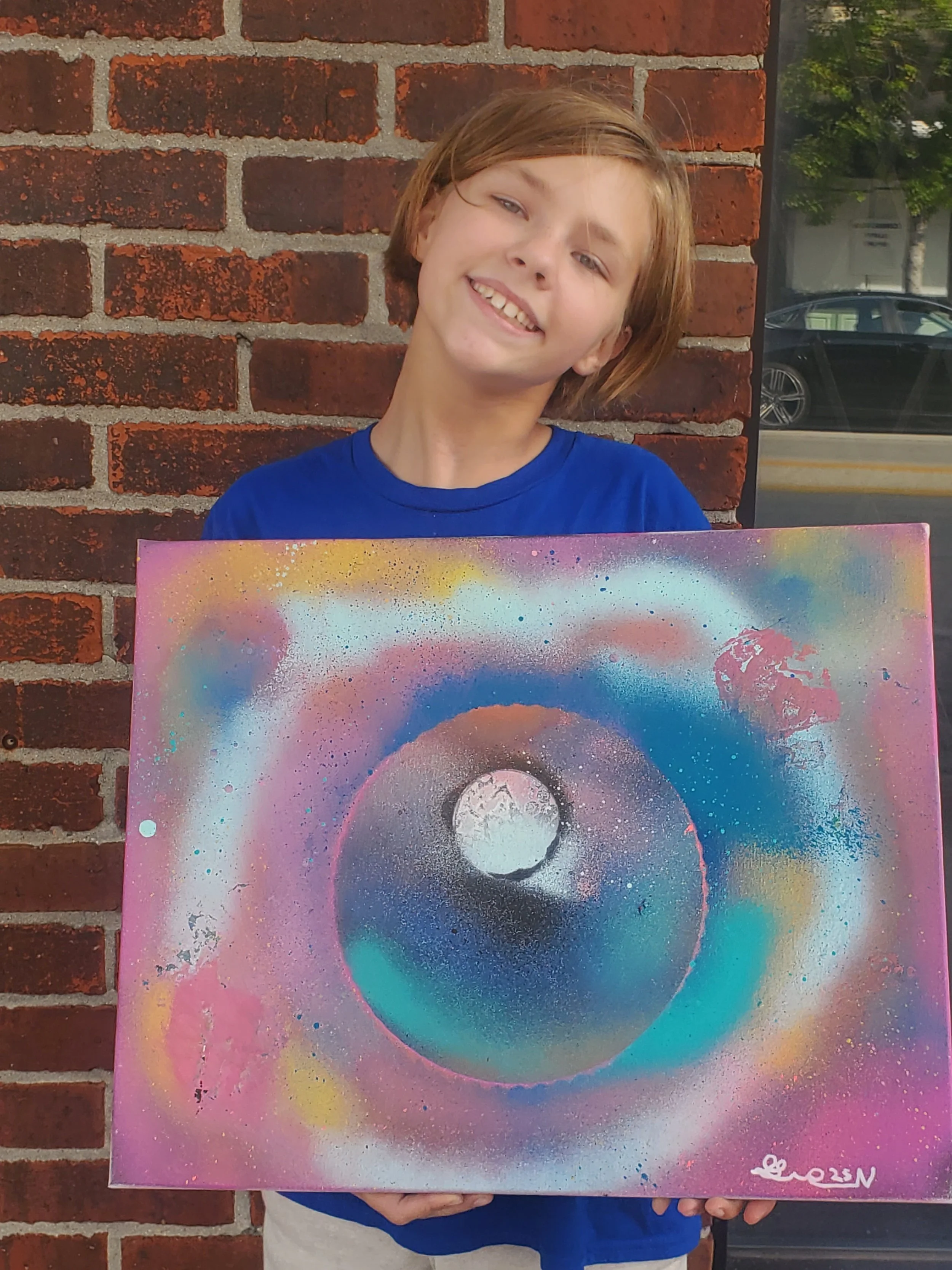 A child with light brown hair, wearing a blue t-shirt, standing outside against a red brick wall, holding a colorful abstract painting featuring a circular design with multiple shades of pink, blue, purple, and white.