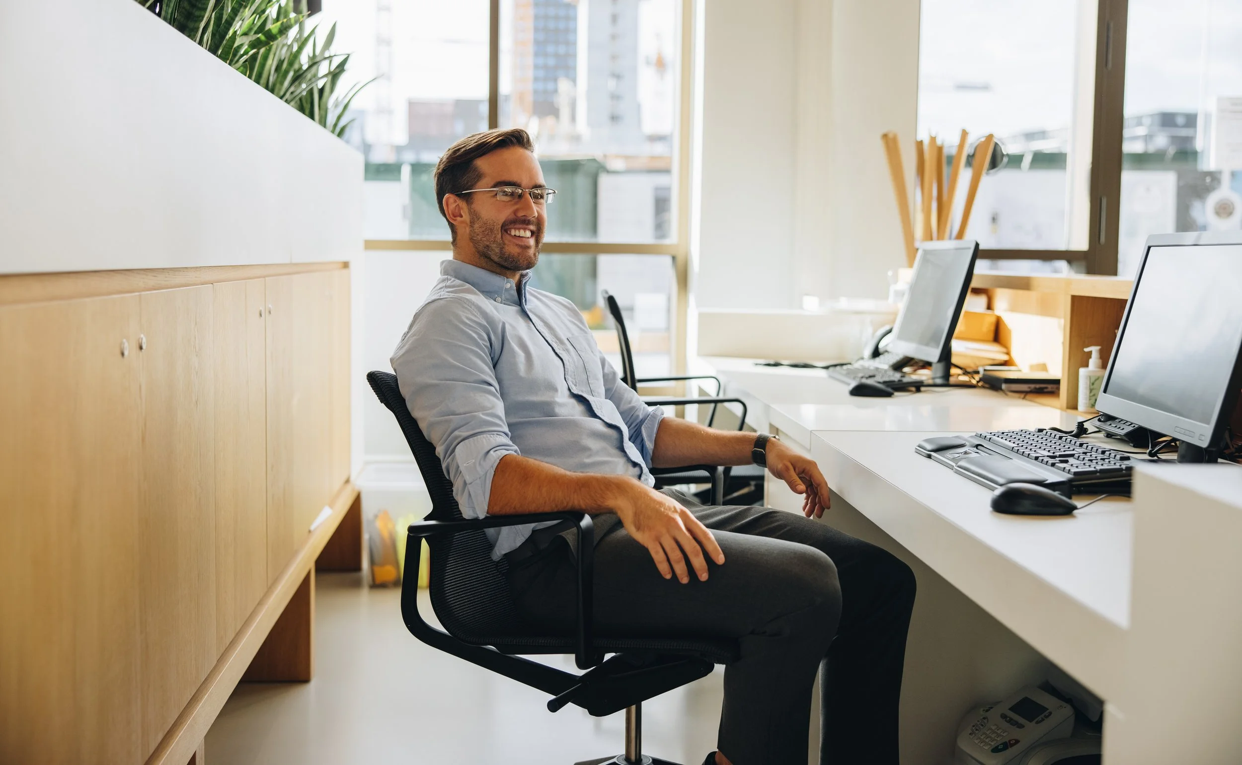 Man sitting at a desk in an office with computer monitors, a keyboard, and a mouse, smiling and looking to the side.