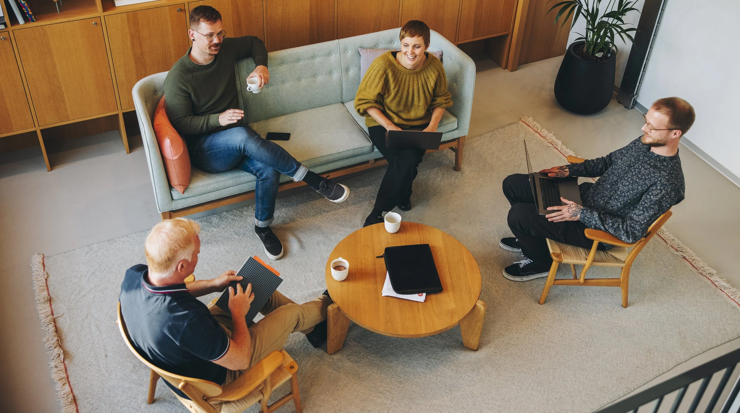 A group of five people sitting in a cozy living room or office space having a discussion. They are sitting around a small round wooden table that holds a black folder, documents, and three cups of coffee or tea. Two of the people are on a sofa, two are on wooden chairs, and one is sitting cross-legged on the floor near the sofa, all engaged in conversation and smiling.