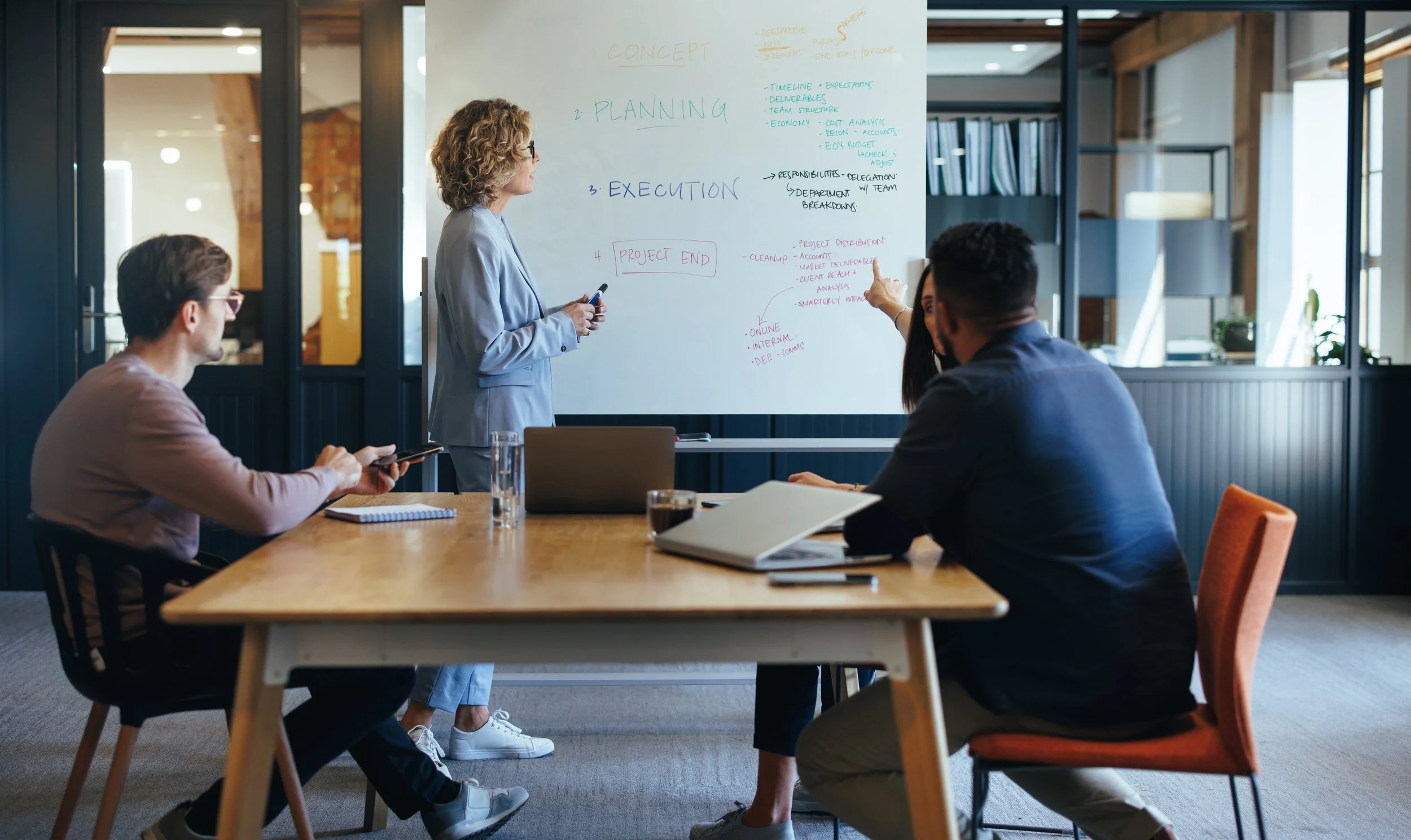 A woman in a blue suit leading a meeting in a modern office conference room, standing next to a whiteboard with handwritten notes about project planning, while three colleagues listen and take notes around a wooden table.